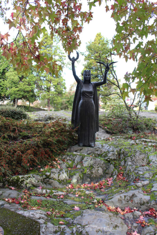 Bronze statue on a rocky hill of a woman wearing a crown and holding a laurel wreath and a bird in up-stretched arms