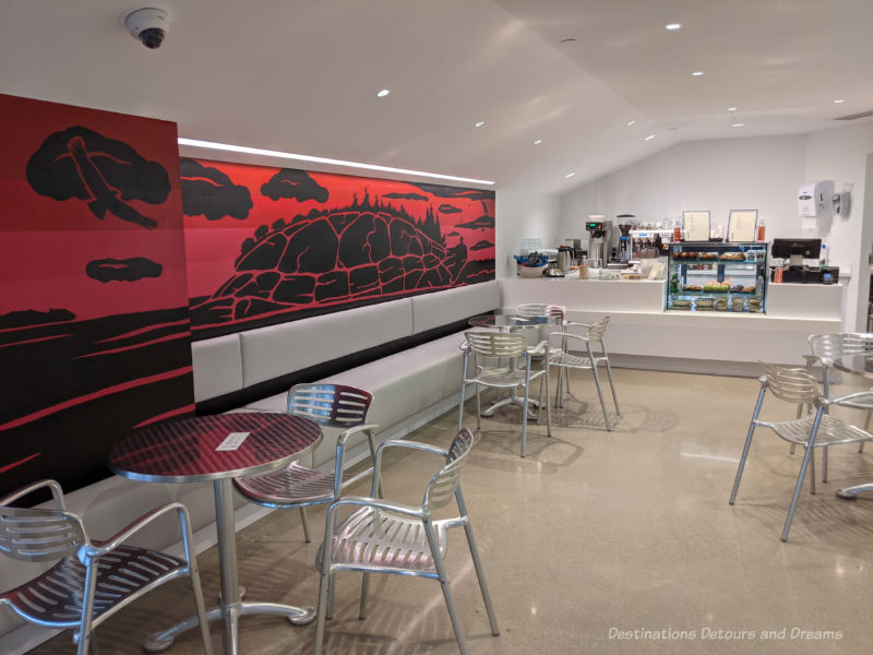 Café at Qaumajuq with food counter, chrome round tables and chairs, and a red and black Inuit art piece along one wallQau