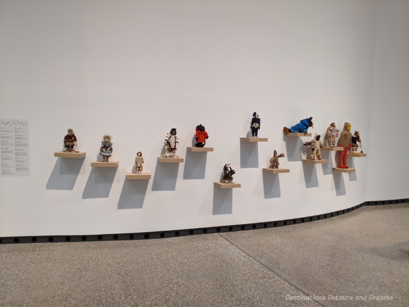 Collection of Inuit dolls displayed on individual wood shelves on the side of large white wall at Qaumajq