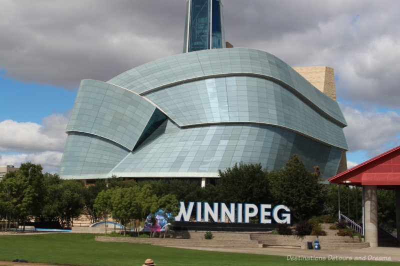 Winnipeg sign on the lawn in front of the curved glass wings of the Canadian Museum for Human Rights in Winnipeg, Manitoba