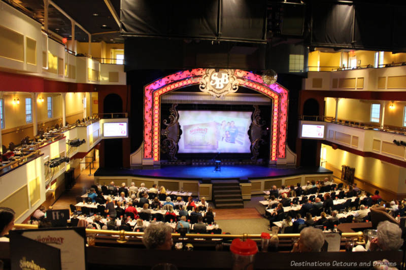 Long dinner tables in front of a raised stage on the main floor of the Showboat Branson Belle area with balcony areas along the side