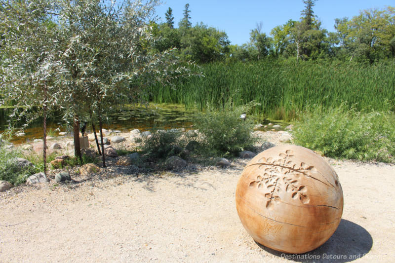 Large carved wooden ball on gravelled are in front of pond surrounded by tall grasses and shrubs