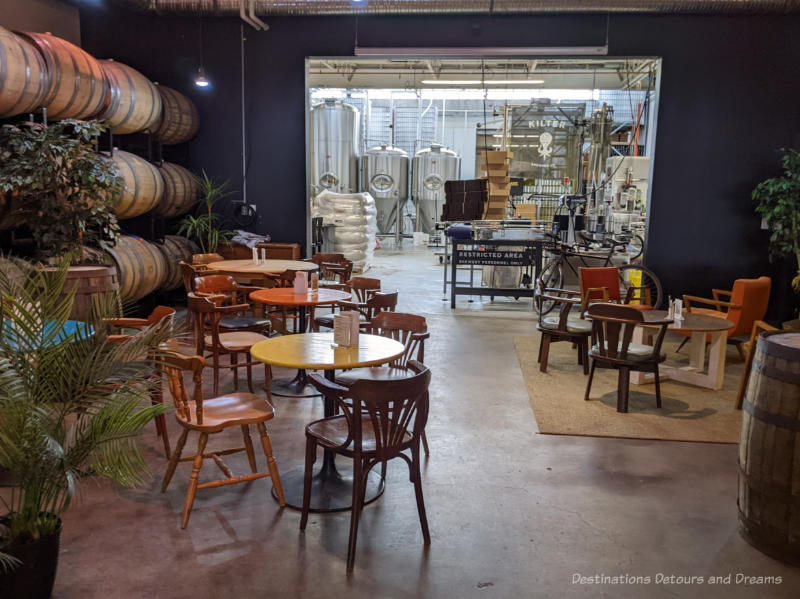 Kilter Brewing Taproom and Brewery Round tables and chairs in the Winnipeg craft brewery Kilter Brewing taproom with wooden kegs of beer along one wall and an opening in the wall showing the brewery in the back