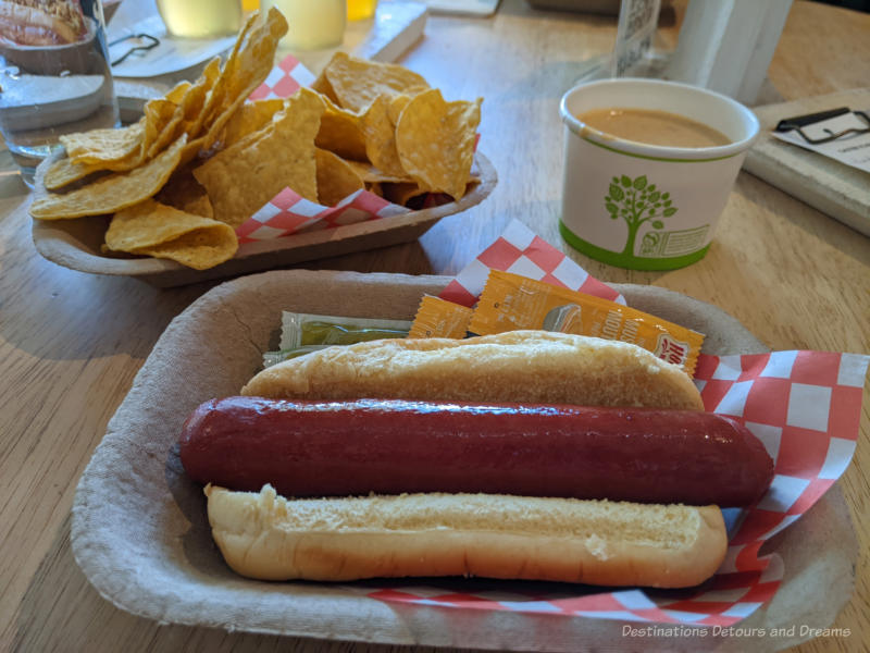 Plain hot dog on a cardboard plate with a tray of tortilla chips and bowl of melted cheese in the background