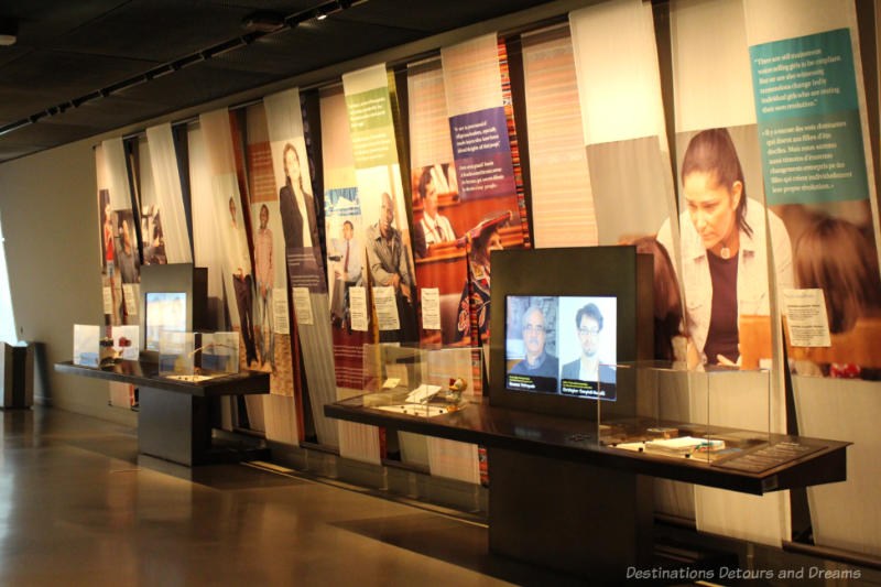 Display wall at CMHR contain placards, written information, digital screens, and artifacts under glass about human rights defenders