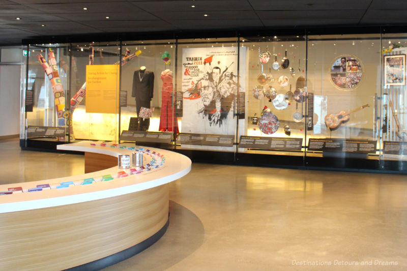 A wall of exhibits behind glass and a circular table in centre of CMHR Inspiring Change gallery where people can leave comments